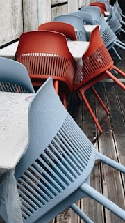 A row of modern chairs, some in blue and some in red, are aligned next to tables with white tablecloths. The chairs have a sleek design with slatted backs and are placed on a wooden deck. Water droplets are visible on the chairs, indicating recent rain.