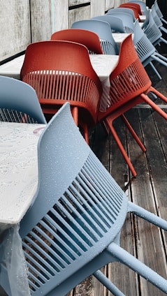 A row of modern chairs, some in blue and some in red, are aligned next to tables with white tablecloths. The chairs have a sleek design with slatted backs and are placed on a wooden deck. Water droplets are visible on the chairs, indicating recent rain.