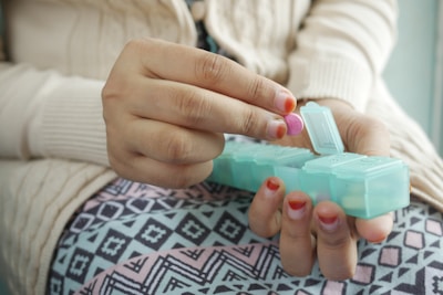A person holding a pink pill above a turquoise pill organizer, which is partially open. The person is wearing a beige knitted sweater and clothes with a geometric pattern.