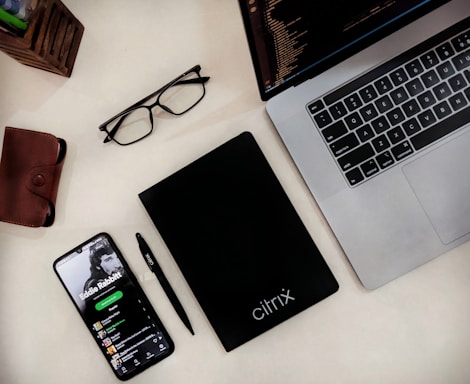 A workspace setup featuring a laptop with a visible keyboard and part of its screen showing code. A smartphone displaying a music app is placed nearby, along with a black notebook labeled 'citrix'. Additional items include a brown leather wallet, a pair of black glasses, and a black pen. A wooden pen holder with pens is partly visible.