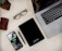 A workspace setup featuring a laptop with a visible keyboard and part of its screen showing code. A smartphone displaying a music app is placed nearby, along with a black notebook labeled 'citrix'. Additional items include a brown leather wallet, a pair of black glasses, and a black pen. A wooden pen holder with pens is partly visible.
