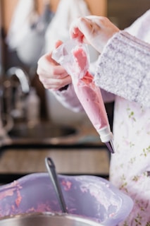 A person is holding a piping bag filled with pink icing, preparing to decorate. The hands are gently squeezing the bag, and a mixing bowl with pink icing is visible in the foreground.