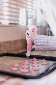 A person is using a piping bag to create pink heart-shaped pastries on a baking tray. The scene is set in a kitchen with soft, natural lighting coming through window blinds. The focus is on the hand applying the dough, which is neatly arranged on a patterned baking sheet.