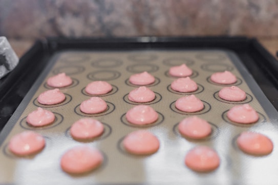 Several small pink meringue-like drops are placed evenly on a silicone baking mat, which is on a baking sheet. The background appears to be a kitchen countertop.