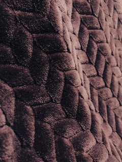 A close-up of a textured surface with a woven pattern made of soft, plush material. The fabric appears lush and densely packed, exhibiting a geometric design with intersecting shapes.