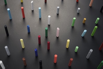 A variety of colorful chalk sticks are arranged upright on a dark surface, creating a contrast between the bright hues and the background. The chalk sticks are evenly spaced in a grid-like pattern, showcasing an array of colors including blue, green, red, yellow, and white.