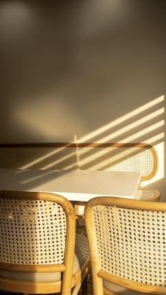A minimalist setting featuring a white table with two woven-back chairs. Sunlight creates a pattern of stripes on the neutral walls, contributing to the warm atmosphere.