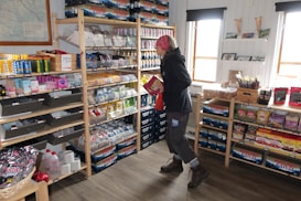 A small shop or store with shelves stocked with various grocery items such as snacks, drinks, and household supplies. A person is browsing the shelves and carrying packages, wearing a jacket, pants, and boots. The store is well-lit with natural light coming through the windows, and there is a wall map visible in the background.