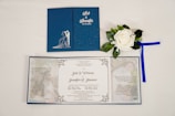 A wedding invitation set displayed on a white surface. The invitation features a dark blue cover with an embossed silhouette of a couple, along with decorative floral patterns. Inside, there is a white panel with printed text detailing the marriage of two individuals. Accompanying the invitation is a white artificial rose with green leaves and a blue ribbon.