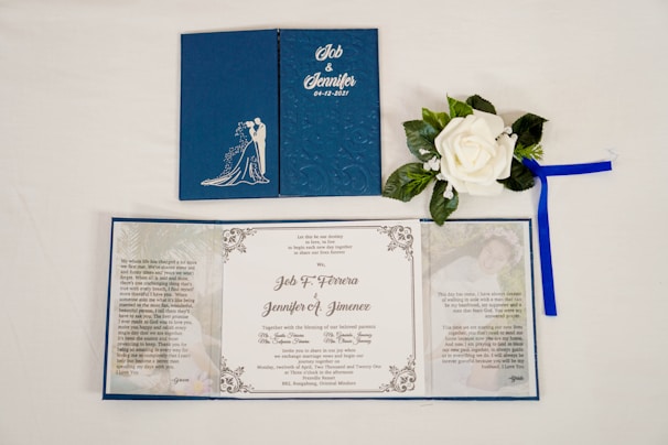 A wedding invitation set displayed on a white surface. The invitation features a dark blue cover with an embossed silhouette of a couple, along with decorative floral patterns. Inside, there is a white panel with printed text detailing the marriage of two individuals. Accompanying the invitation is a white artificial rose with green leaves and a blue ribbon.
