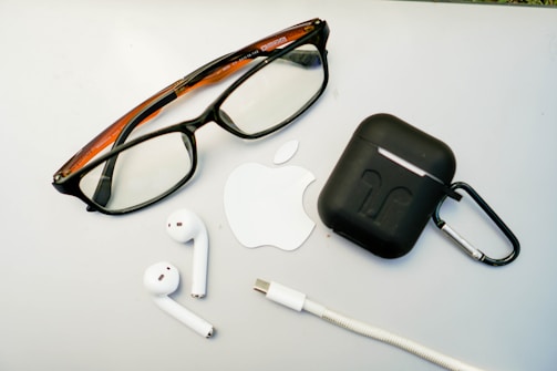 A pair of glasses, wireless earbuds, a charging case, a USB-C cable, and an Apple logo sticker are arranged on a light surface.