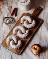 Three powdered pastries are arranged on a wooden board, accompanied by a glass of milk, cinnamon sticks, a sieve with powdered sugar, and an apple. The setting is on a textured fabric surface.