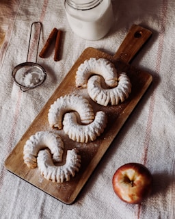 Three powdered pastries are arranged on a wooden board, accompanied by a glass of milk, cinnamon sticks, a sieve with powdered sugar, and an apple. The setting is on a textured fabric surface.