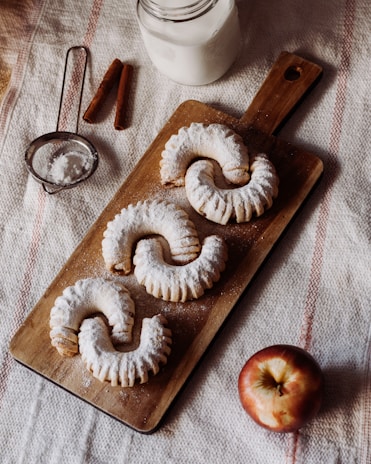 Three powdered pastries are arranged on a wooden board, accompanied by a glass of milk, cinnamon sticks, a sieve with powdered sugar, and an apple. The setting is on a textured fabric surface.