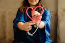 A person wearing a blue garment is holding a pink stethoscope shaped into a heart. The focus is on their hands and the stethoscope, suggesting a connection to healthcare or caring professions.