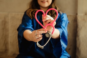 A person wearing a blue garment is holding a pink stethoscope shaped into a heart. The focus is on their hands and the stethoscope, suggesting a connection to healthcare or caring professions.