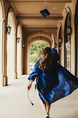 A graduate in a blue gown tossing a cap in a stone archway