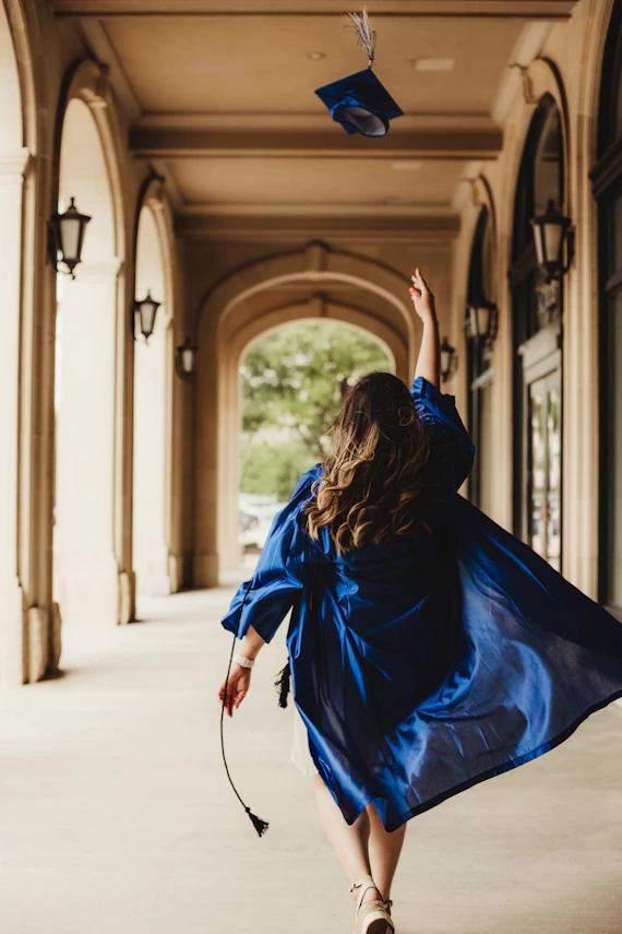 A graduate in a blue gown tossing a cap in a stone archway