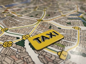 A detailed map with streets and buildings illustrated, featuring a notable focus on a yellow taxi sign placed on top of the map. The streets are marked with numbers and the layout suggests an urban environment with various buildings and greenery.