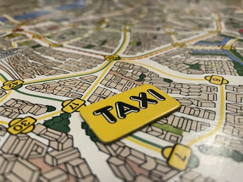 A detailed map with streets and buildings illustrated, featuring a notable focus on a yellow taxi sign placed on top of the map. The streets are marked with numbers and the layout suggests an urban environment with various buildings and greenery.