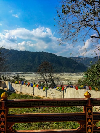 A serene landscape featuring a range of hills covered in greenery under a partly cloudy blue sky. In the foreground, a decorative wooden railing and a string of colorful prayer flags are visible, along with a white wall. Trees with sparse leaves stand on the side, enhancing the tranquil atmosphere.