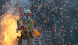 A character in futuristic armor, holding a weapon, stands amidst a dramatic scene filled with glowing embers and smoke. The background is dark, contrasted by bright flames and floating sparks.