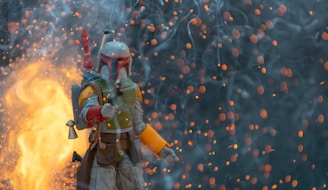 A character in futuristic armor, holding a weapon, stands amidst a dramatic scene filled with glowing embers and smoke. The background is dark, contrasted by bright flames and floating sparks.