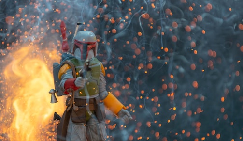 A character in futuristic armor, holding a weapon, stands amidst a dramatic scene filled with glowing embers and smoke. The background is dark, contrasted by bright flames and floating sparks.
