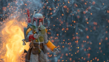A character in futuristic armor, holding a weapon, stands amidst a dramatic scene filled with glowing embers and smoke. The background is dark, contrasted by bright flames and floating sparks.