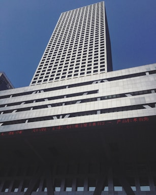 A tall skyscraper with a grid-like pattern of windows against a clear blue sky. The building is modern, with a silver and gray facade, and features an LED ticker displaying text in red.