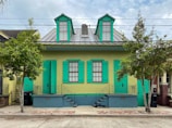 A vibrant house with two turquoise-painted shutters on its windows and a matching door. The house front is light green, with a metal roof featuring two dormers. It is flanked by two trees and has a small front porch with decorative wrought-iron railings. Two staircases lead up to the porch, with street numbers 1018 and 1020 displayed.