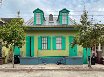 A vibrant house with two turquoise-painted shutters on its windows and a matching door. The house front is light green, with a metal roof featuring two dormers. It is flanked by two trees and has a small front porch with decorative wrought-iron railings. Two staircases lead up to the porch, with street numbers 1018 and 1020 displayed.