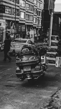 A city street scene with a parked motorcycle in the foreground. The motorcycle has a prominent license plate visible. In the background, a street with several people walking and a few cars is visible. The surrounding buildings are tall and have numerous windows.