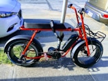 A parked red electric bike with wide tires is secured to a pole on a city street. It is positioned next to two cars and has a sleek, modern design with a sturdy frame and a battery compartment.