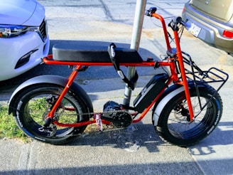 A parked red electric bike with wide tires is secured to a pole on a city street. It is positioned next to two cars and has a sleek, modern design with a sturdy frame and a battery compartment.