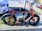A parked red electric bike with wide tires is secured to a pole on a city street. It is positioned next to two cars and has a sleek, modern design with a sturdy frame and a battery compartment.