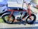 A parked red electric bike with wide tires is secured to a pole on a city street. It is positioned next to two cars and has a sleek, modern design with a sturdy frame and a battery compartment.