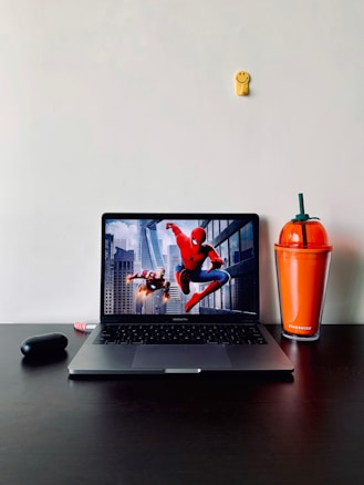 A minimalist desk setup features a laptop displaying an action scene with superheroes against a city backdrop. To the right of the laptop is an orange Starbucks tumbler with a green straw resembling a pumpkin. On the left, there is a black wireless earphone case. A small yellow smiley face decoration is attached to the wall above the laptop.