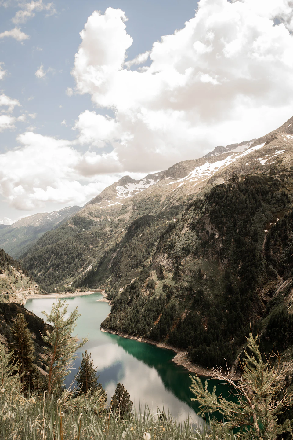 The image depicts a serene mountain landscape with a large lake nestled amidst the mountains, surrounded by lush green trees and shrubs. The sky above is clear blue with fluffy white clouds scattered across it. The perspective of the photo is from a high vantage point, providing an expansive view of the natural beauty below.