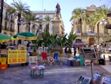 A bustling outdoor marketplace with colorful stalls and umbrellas. A statue stands on a raised platform surrounded by palm trees in the center of a plaza, with historic buildings in the background. Various goods, including fresh produce and magazines, are on display, and people are engaging in conversation and shopping.