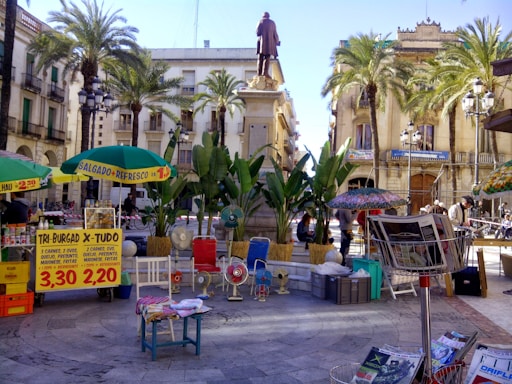 A bustling outdoor marketplace with colorful stalls and umbrellas. A statue stands on a raised platform surrounded by palm trees in the center of a plaza, with historic buildings in the background. Various goods, including fresh produce and magazines, are on display, and people are engaging in conversation and shopping.