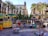 A bustling outdoor marketplace with colorful stalls and umbrellas. A statue stands on a raised platform surrounded by palm trees in the center of a plaza, with historic buildings in the background. Various goods, including fresh produce and magazines, are on display, and people are engaging in conversation and shopping.