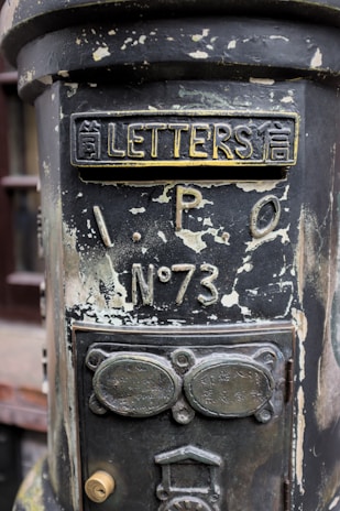 An old, weathered postbox with peeling paint and ornate metalwork. The top of the box features the word 'LETTERS' alongside Asian characters. Below, there is an inscription 'I.P.O No 73'. The surface appears corroded, giving it a vintage and historical appearance. The lower section has additional metal plaques with inscriptions and a keyhole lock.