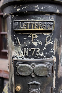 An old, weathered postbox with peeling paint and ornate metalwork. The top of the box features the word 'LETTERS' alongside Asian characters. Below, there is an inscription 'I.P.O No 73'. The surface appears corroded, giving it a vintage and historical appearance. The lower section has additional metal plaques with inscriptions and a keyhole lock.