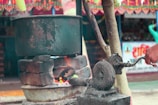 A large metal pot is placed atop a makeshift stove constructed with bricks. A visible fire burns below the pot, suggesting cooking or boiling. The scene includes a traditional manual blower used to keep the fire alive. The background features colorful decorations and a partially visible poster with text.