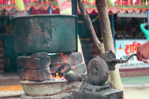 A large metal pot is placed atop a makeshift stove constructed with bricks. A visible fire burns below the pot, suggesting cooking or boiling. The scene includes a traditional manual blower used to keep the fire alive. The background features colorful decorations and a partially visible poster with text.