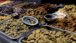 A variety of dried fruits and sweets are displayed in metal trays on a market stall. The assortment includes a range of textures and colors, with some items covered in spices or powders. Small bowls are placed amidst the trays, likely for sampling or additional organization.