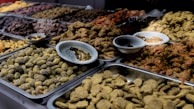 A variety of dried fruits and sweets are displayed in metal trays on a market stall. The assortment includes a range of textures and colors, with some items covered in spices or powders. Small bowls are placed amidst the trays, likely for sampling or additional organization.