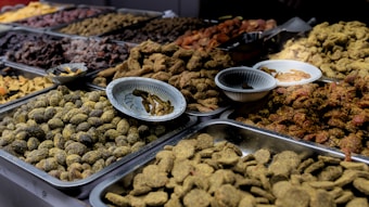 A variety of dried fruits and sweets are displayed in metal trays on a market stall. The assortment includes a range of textures and colors, with some items covered in spices or powders. Small bowls are placed amidst the trays, likely for sampling or additional organization.