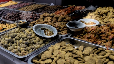 A variety of dried fruits and sweets are displayed in metal trays on a market stall. The assortment includes a range of textures and colors, with some items covered in spices or powders. Small bowls are placed amidst the trays, likely for sampling or additional organization.